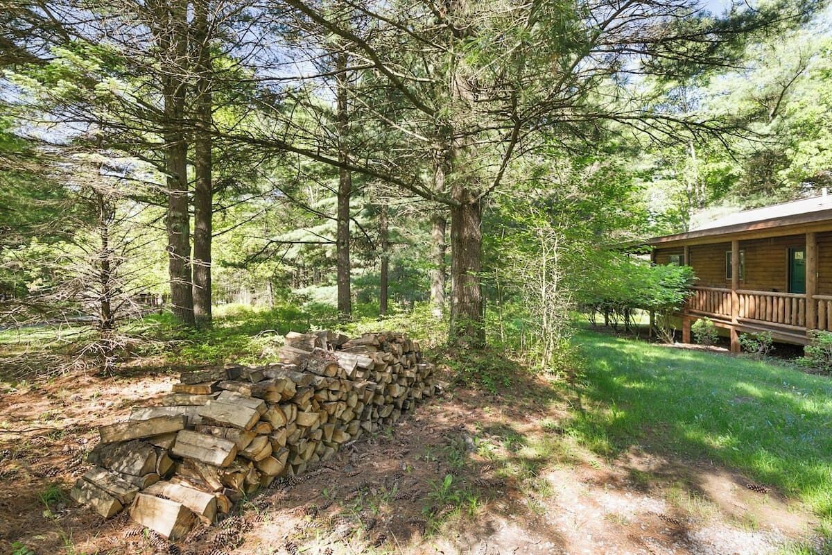 A cabin surrounded by trees, with a stack of firewood and a grassy area in front.
