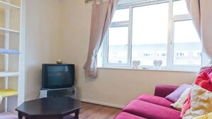 Ein Zimmer mit Fernseher auf einem Schrank, einem rosa Sofa, einem kleinen Tisch und einem Fenster mit Vorhängen.