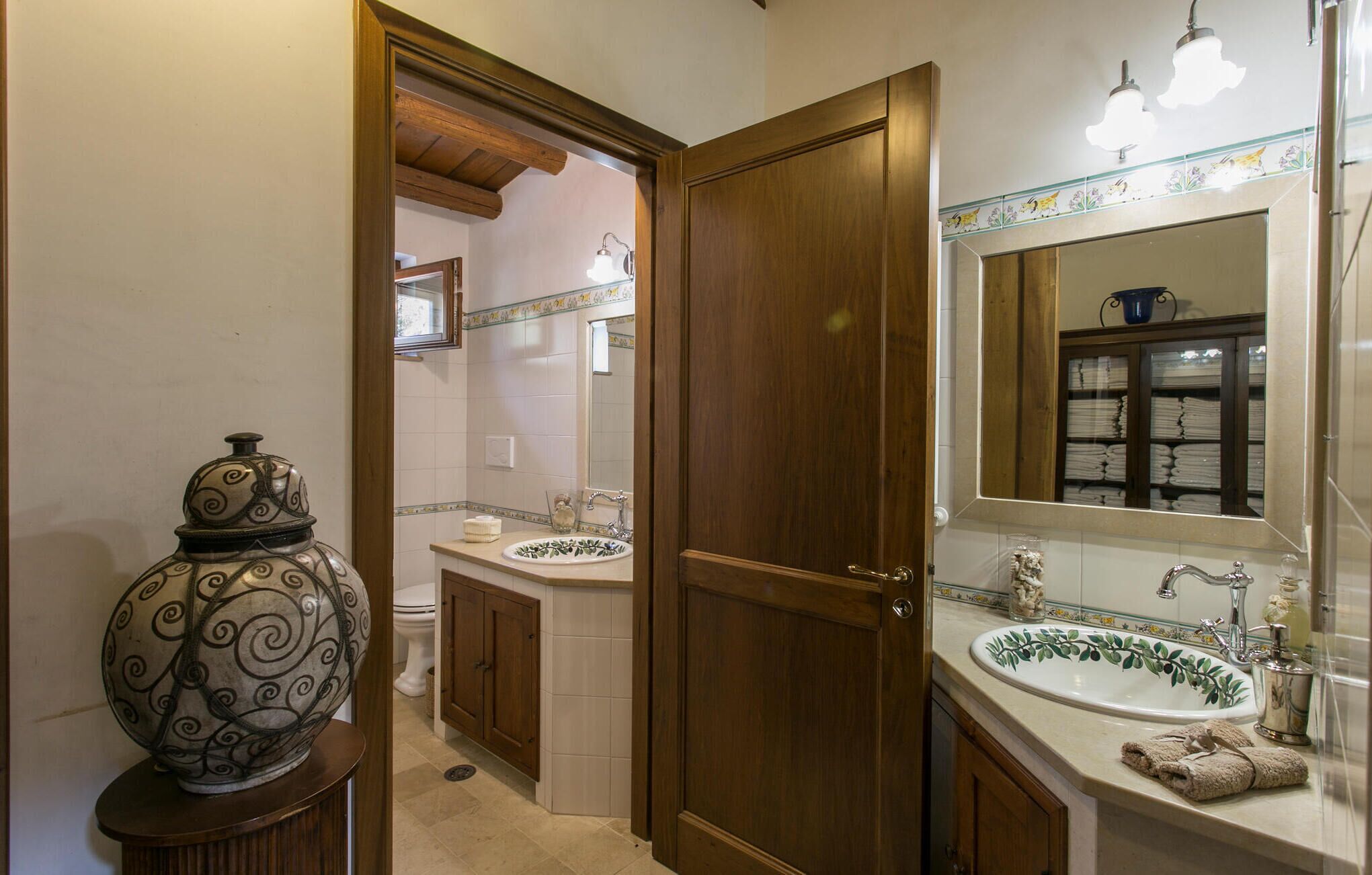 A bathroom with a wooden door, two sinks with decorative tiles, a mirror with a towel rack, and a decorative vase on a pedestal.