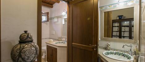 A bathroom with a wooden door, two sinks with decorative tiles, a mirror with a towel rack, and a decorative vase on a pedestal.
