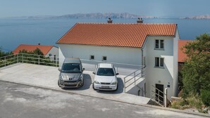 Un edificio de dos plantas con techo de tejas rojas, dos coches aparcados y vistas al mar y las montañas al fondo.