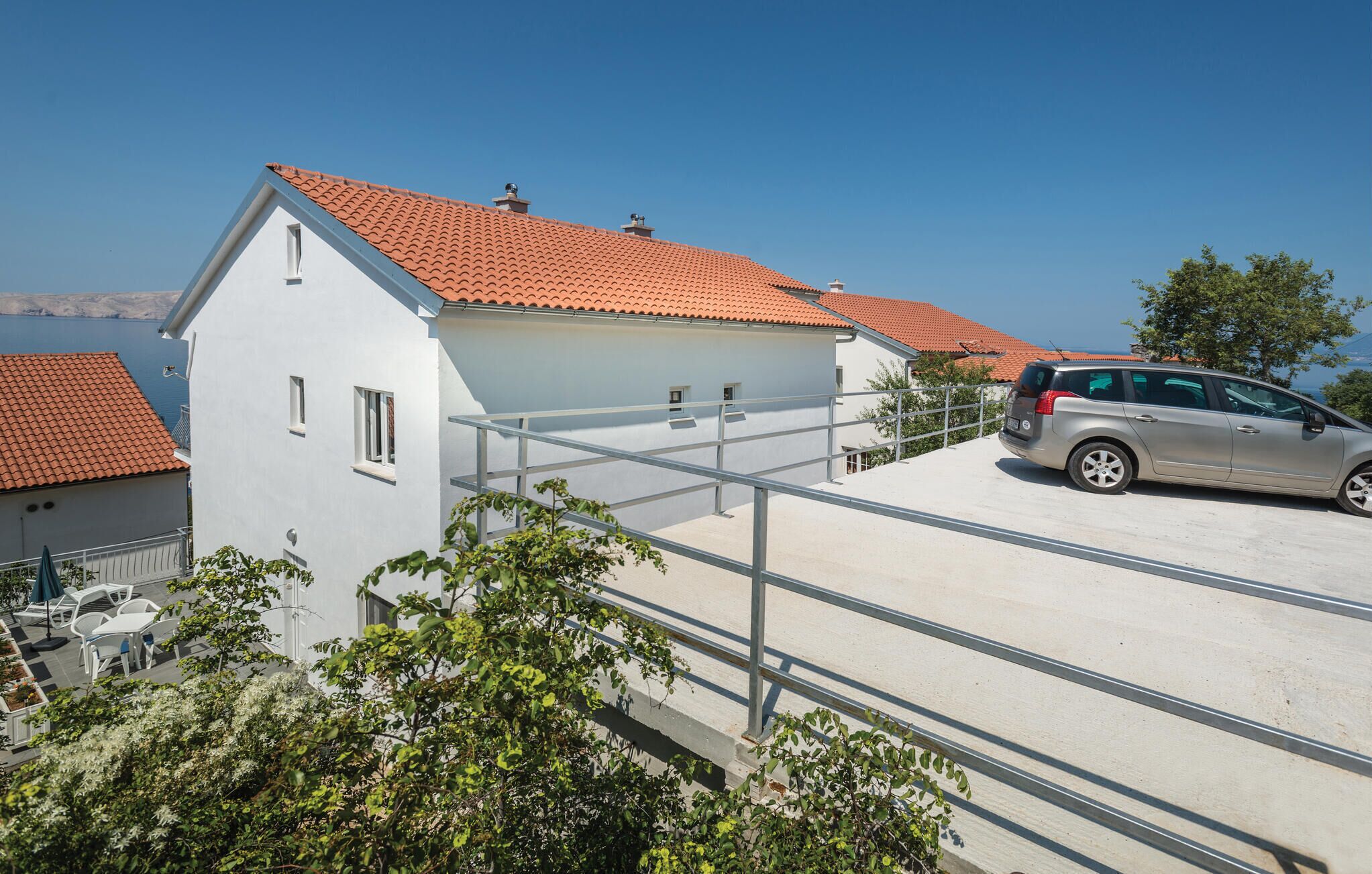 A white building with a red-tiled roof, a car parked in front, and a balcony with metal railings.