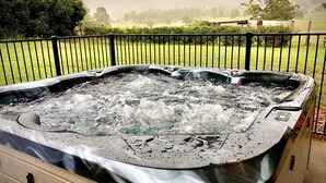 A hot tub with a wooden deck, surrounded by a metal fence, with a grassy field and trees in the background. - In the Hunter Valley and need a place to stay for a big group? Look no further (Bellbird)