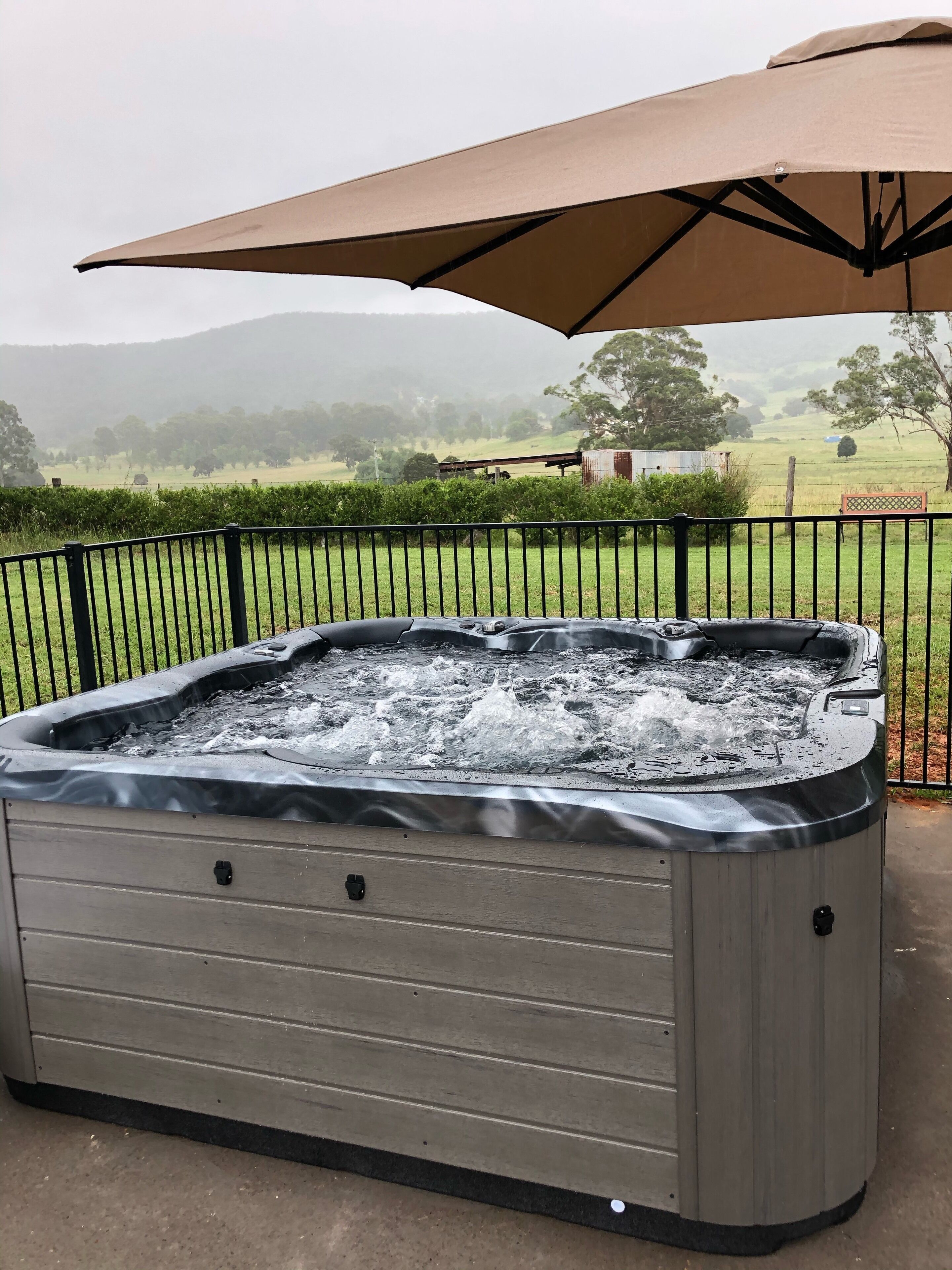Ein Whirlpool mit Metallrahmen und einer beigen Abdeckung, draußen aufgestellt mit Blick auf eine grüne Landschaft und Hügel im Hintergrund.