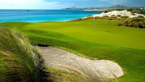 A golf course with a sand bunker, a flagstick, and a view of the ocean and distant hills.