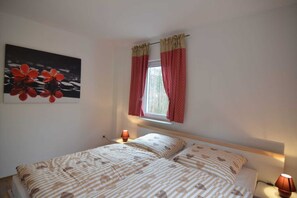 A bedroom with a bed, two bedside lamps, a window with red curtains, and a wall-mounted painting of red flowers.