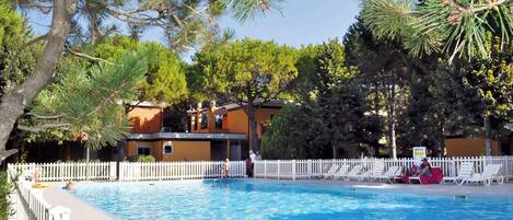 A swimming pool with a white fence, surrounded by trees and a building with a white picket fence.