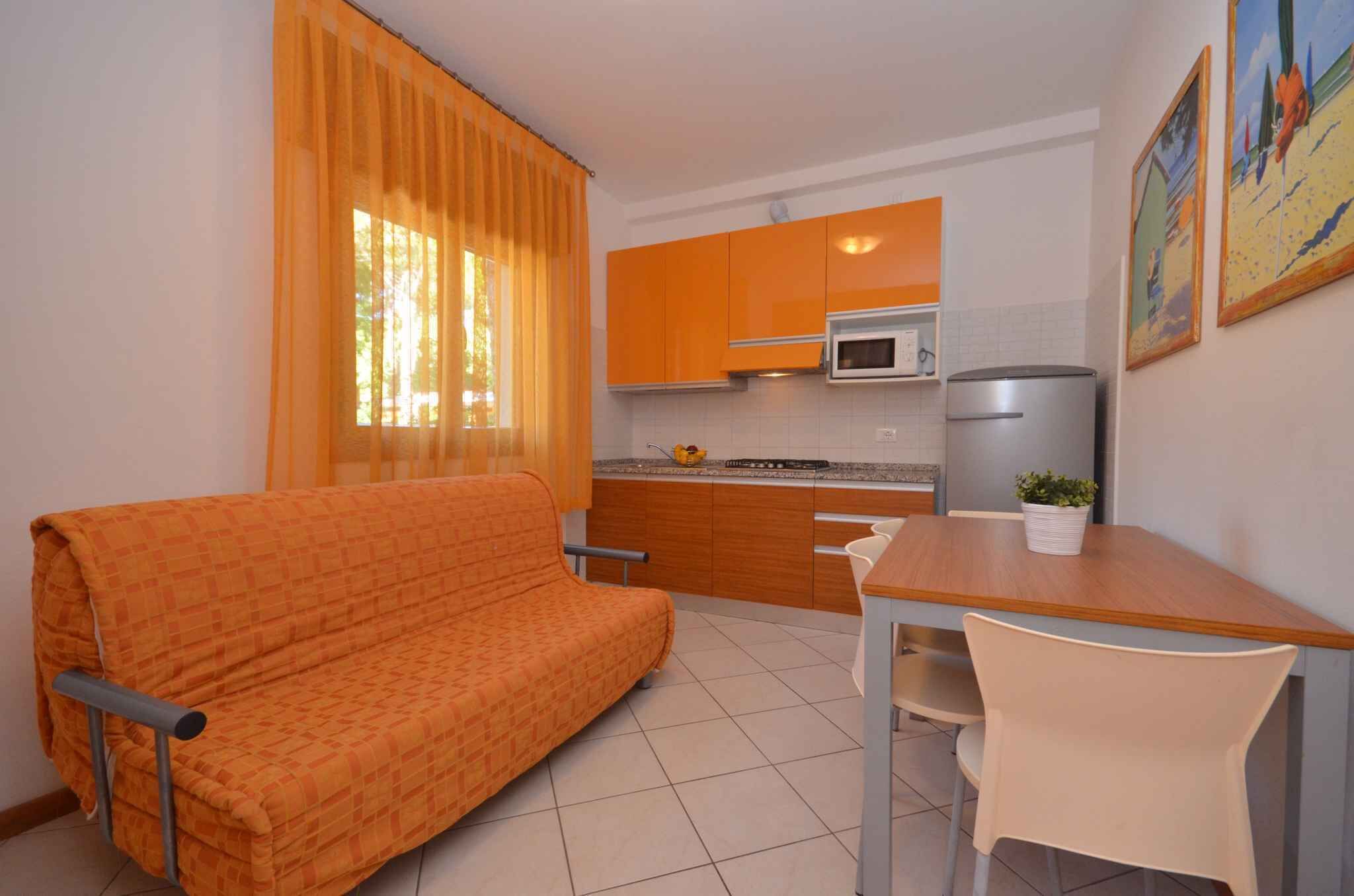 A compact kitchen with orange cabinets, a microwave, and a refrigerator. There's a dining table with a chair and a sofa in the living area.