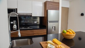 A modern kitchen with a stainless steel refrigerator, a built-in oven, a sink, and a wooden countertop with cheese and fruit.