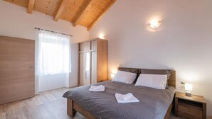 Une chambre avec un plafond en bois, un lit recouvert d’une couverture à motifs gris, deux oreillers blancs et deux serviettes pliées posées sur le lit. On y trouve également une armoire en bois, une fenêtre avec des rideaux légers et une table de chevet avec une lampe.
