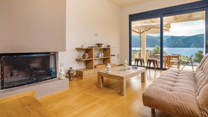 A modern living room with a fireplace, wooden furniture, and a large window offering a view of a lake and mountains. - Seafront Holiday Villa (Skroponeria Politeia)