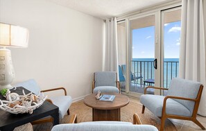 Quarto de hotel com uma mesa redonda de madeira, duas poltronas azul claro, uma mesinha lateral com objetos decorativos e vista para o oceano através de uma porta de vidro deslizante.