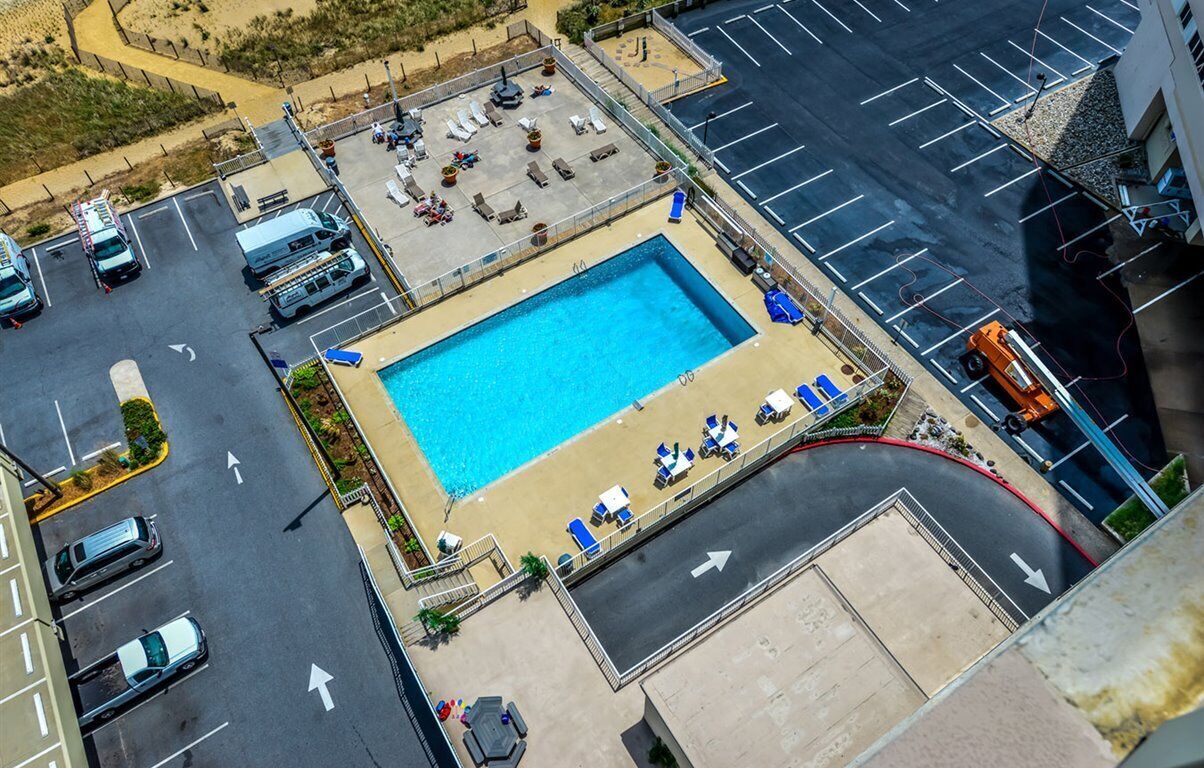 Eine Vogelperspektive zeigt ein Hotel mit Schwimmbad, umgeben von Liegestühlen und Sonnenschirmen, einen Parkplatz und ein Gebäude mit geneigtem Dach.