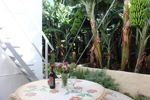 Una mesa con mantel floral, una botella y dos vasos, frente a un fondo de plantas de plátano y una estructura blanca.