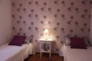 Una habitación con dos camas, una mesita de noche con lámpara y un papel tapiz estampado.