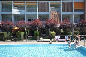 A swimming pool with lounge chairs and a building with multiple balconies in the background. - Holiday flat with air conditioning and balcony (Bibione)