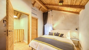 Une chambre avec un plafond en bois, un lit recouvert d’une courtepointe blanche et grise, une table de chevet avec une lampe et une porte en bois.