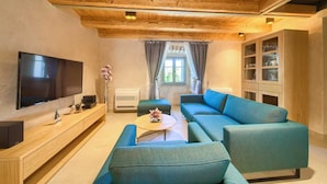 Un salon doté d’un plafond en bois, d’un téléviseur à écran plat, d’un ensemble de canapés, d’une table basse, d’un meuble avec des portes en verre et d’une fenêtre avec des rideaux.