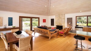 A spacious living area with a wooden ceiling, a stone fireplace, a dining table with wicker chairs, and a kitchen area with a large window. - Yallingup Retreat - *Romantic, overlooking a lake, secluded* (YALLINGUP)