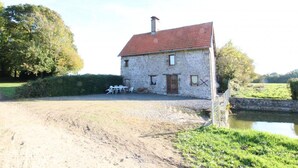 Une maison en pierre avec un toit rouge, une allée en gravier et un petit étang au premier plan.