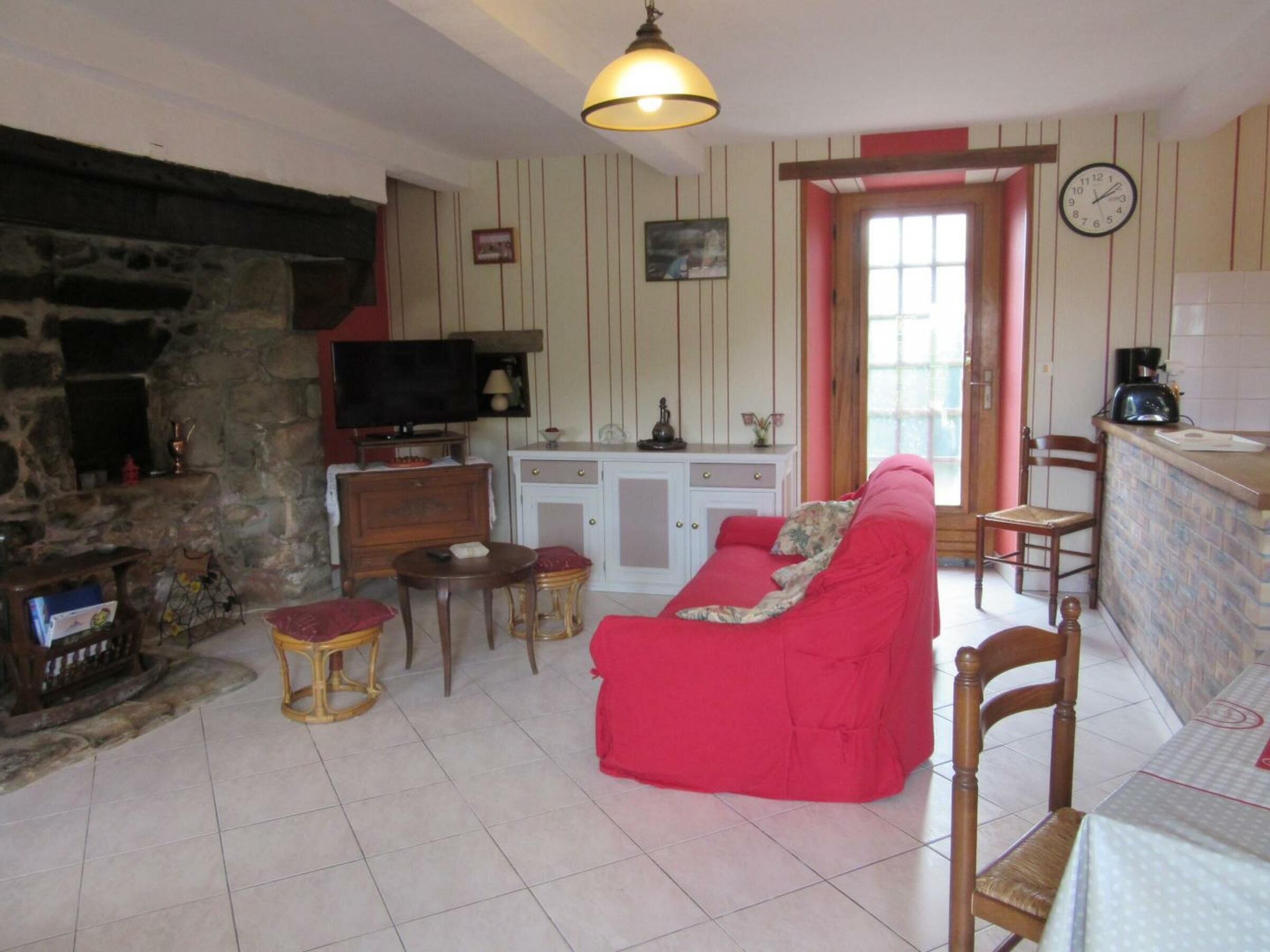 A living room with a stone fireplace, a television, a red sofa, a small table, and a kitchen area with a sink and a clock.