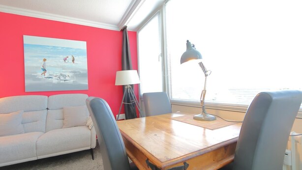 Une pièce avec un mur rouge, un canapé blanc, une table en bois avec des chaises, une lampe de bureau et une fenêtre avec des stores.