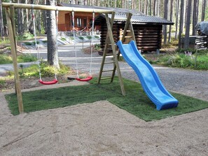 A playground with a slide and swings, surrounded by a wooden cabin and trees.