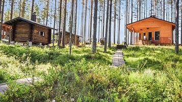 Una cabaña de madera, rodeada de altos árboles y un área con césped, con un camino de madera que la lleva al frente.
