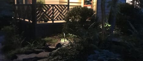 Una cabaña acogedora con terraza de madera y un pequeño porche, rodeada de abundante vegetación y un camino de piedra.