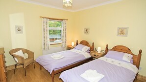 Une pièce avec deux lits en bois, une chaise en osier, une fenêtre avec des rideaux et un luminaire au plafond.