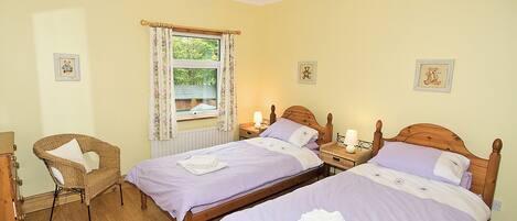 A room with two wooden beds, a wicker chair, a window with curtains, and a ceiling light fixture.