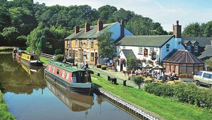 Une ville riveraine où une péniche est amarrée le long d’un canal, avec des bâtiments traditionnels et une promenade au bord de l’eau où l’on peut voir des gens se promener.