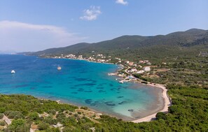 An aerial view of a coastal town with clear turquoise waters, a sandy beach, and lush green hills in the background.