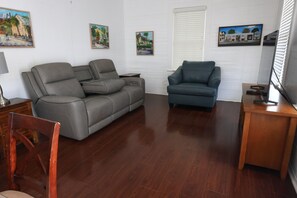 A living room with a grey sofa, a blue armchair, wooden flooring, and wall-mounted paintings. - Heart of Downtown Key West Cottage With Off-Street Parking (Key West)