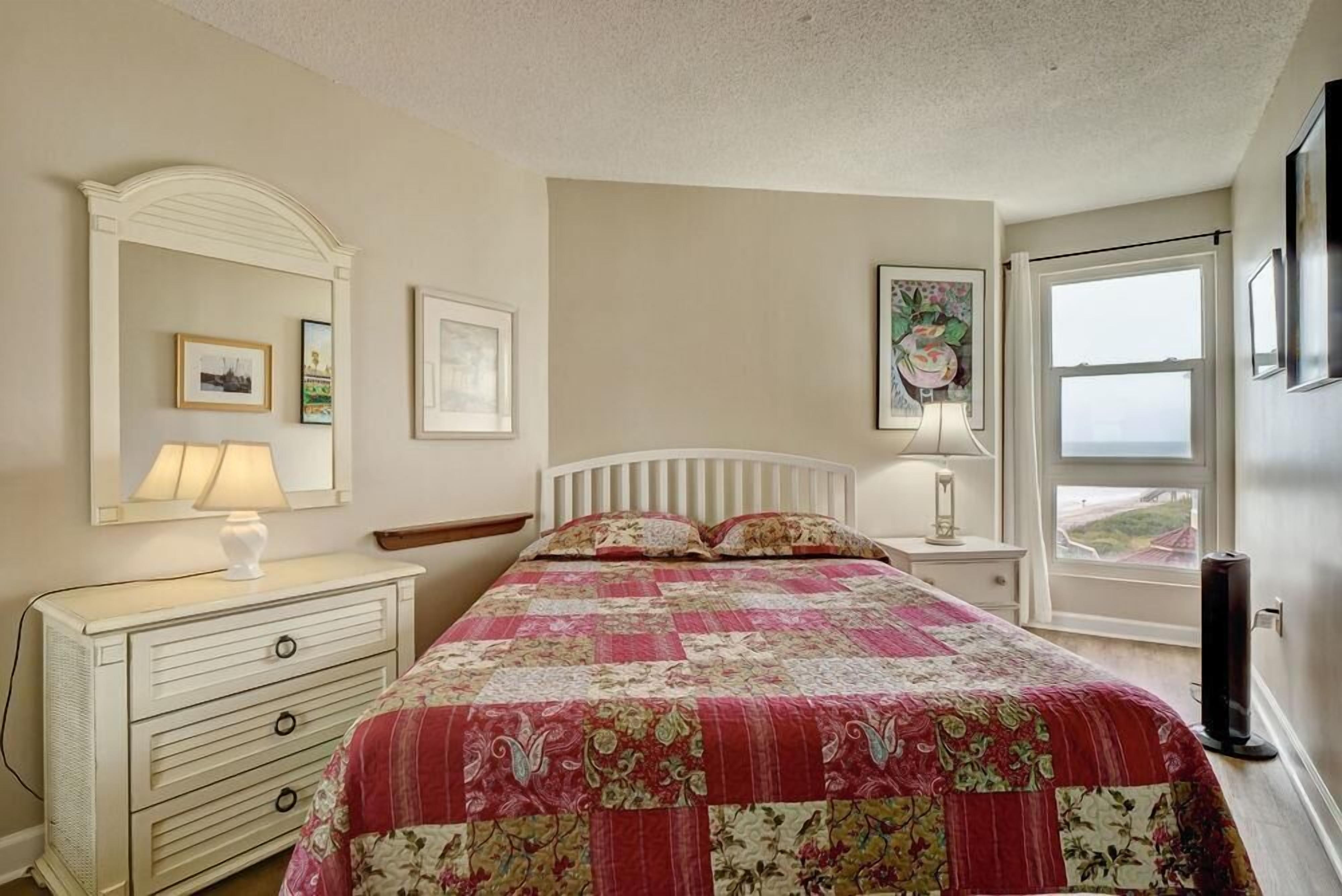 A bedroom with a bed, a dresser, a nightstand, a lamp, a window, and framed pictures on the wall.