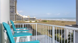 A balcony with turquoise chairs overlooking a beach and a modern building with a curved facade. - Lovely 4th Floor Condo at the Sapphire! Resort Pool, Hot Tub, Spa, Movie Theater & More! (South Padre Island)
