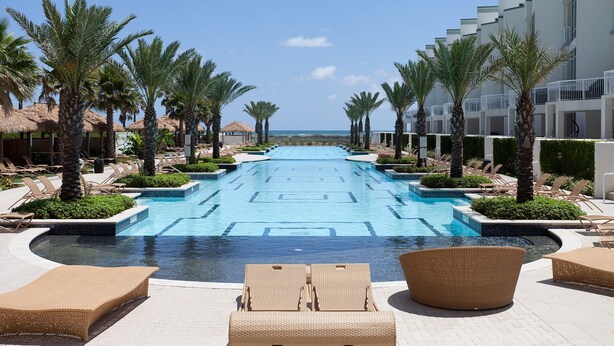A pool area with lounge chairs and palm trees, leading to a beach. - Lovely 4th Floor Condo at the Sapphire! Resort Pool, Hot Tub, Spa, Movie Theater & More! (South Padre Island)
