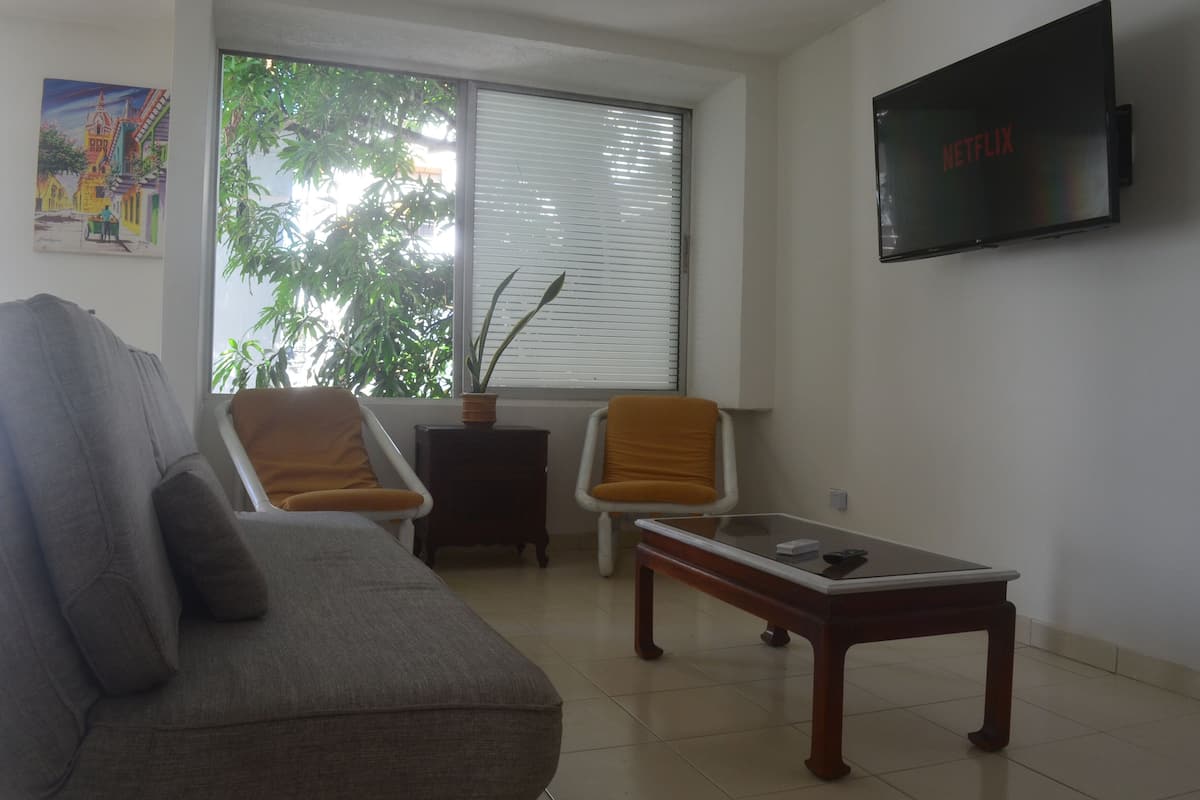 A living room with a sofa, two chairs, a coffee table, a TV mounted on the wall, and a window with blinds.