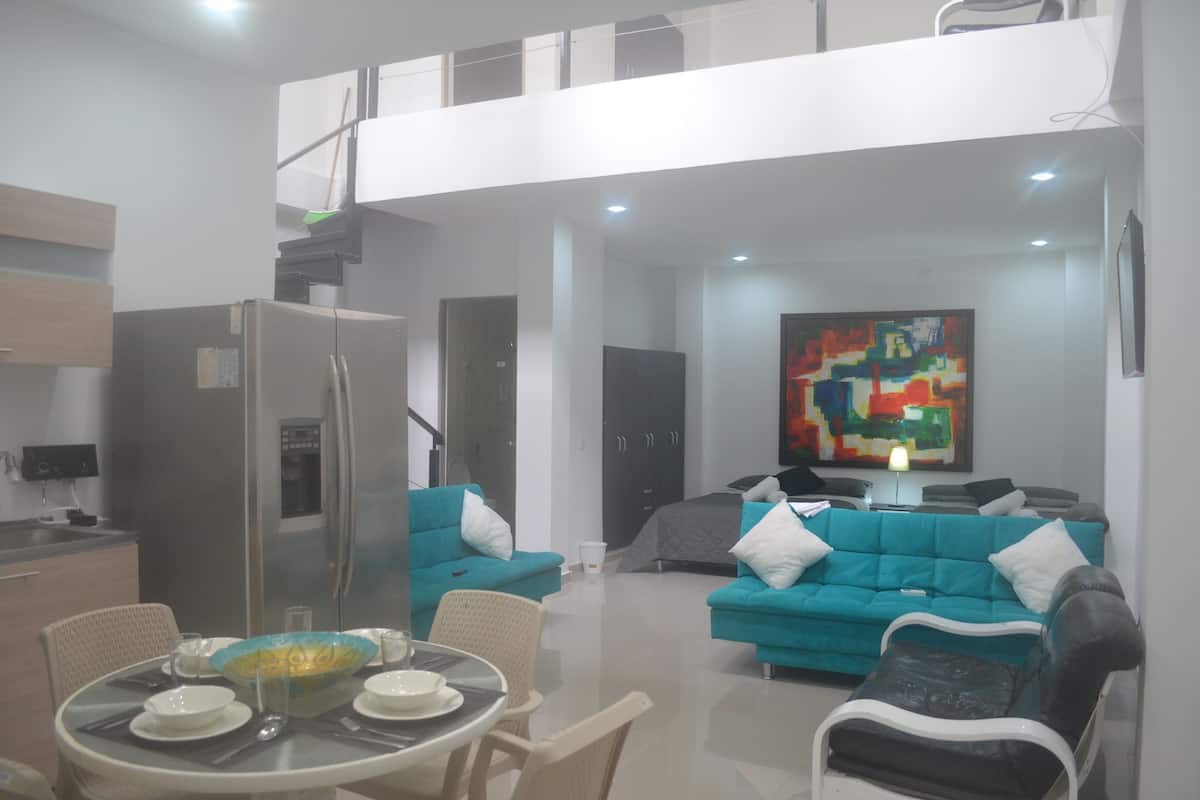 A modern living room with teal sofas, a dining table with chairs, a kitchen area with a refrigerator, and a colorful abstract painting on the wall.