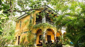 A yellow colonial-style building with a balcony, surrounded by lush greenery and a well-maintained garden.
