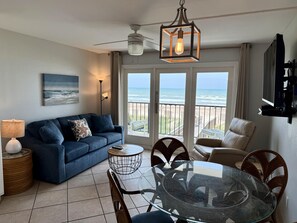 Un soggiorno con divano, tavolo da pranzo con piano in vetro e sedie, una ventola a soffitto e vista sull'oceano attraverso ampie finestre.