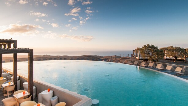 An infinity pool with a view of the ocean and hills, surrounded by lounge chairs and tables.