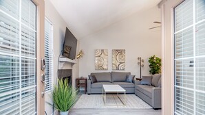 Ein modernes Wohnzimmer mit einem grauen Sofa, einem weißen Couchtisch und einem Kamin. Zwei Bilder hängen an der Wand und es gibt einen Blumentopf.