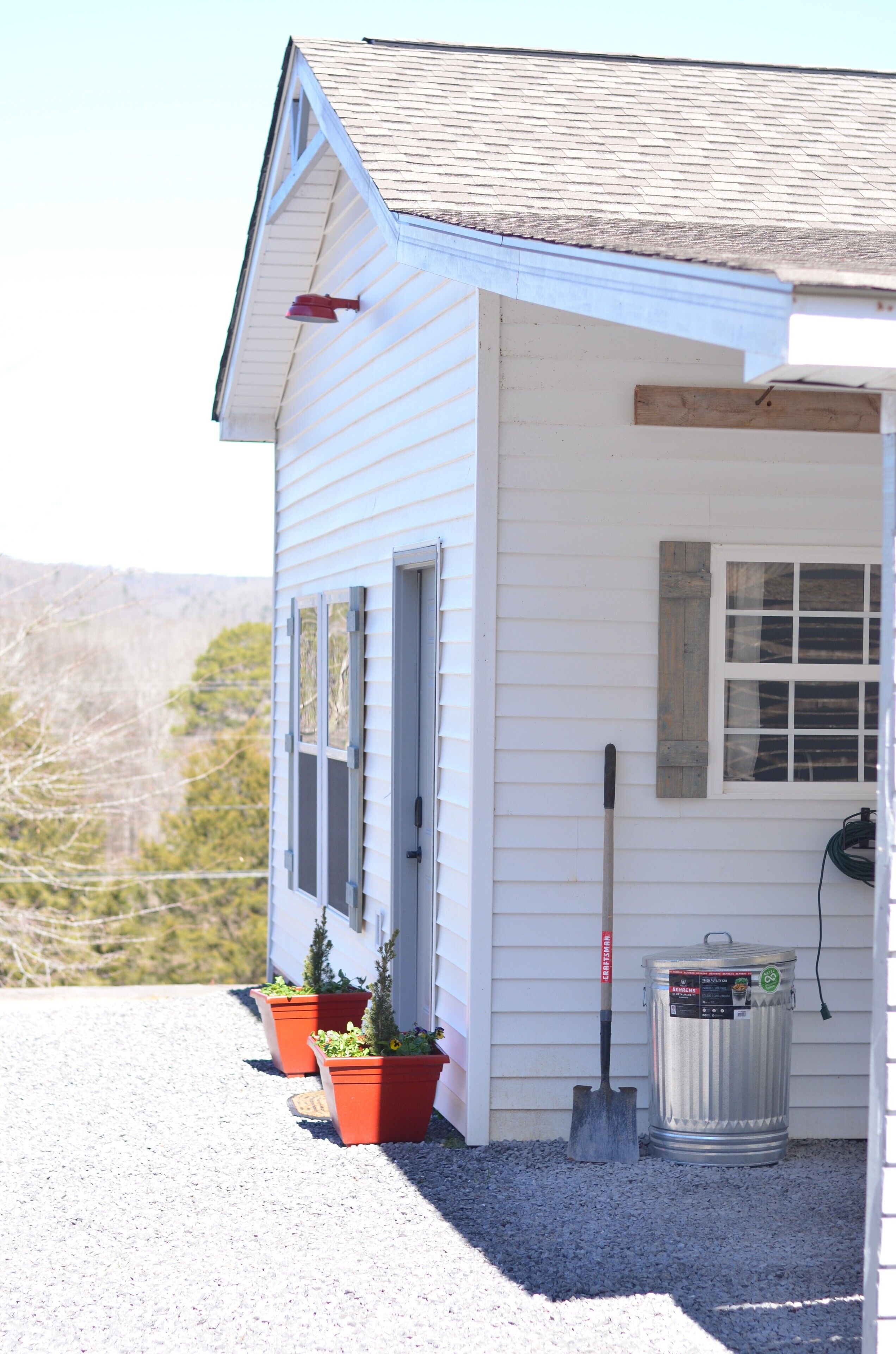 A white building with a sloped roof, a window, and a door. There are two potted plants and a shovel on the ground.