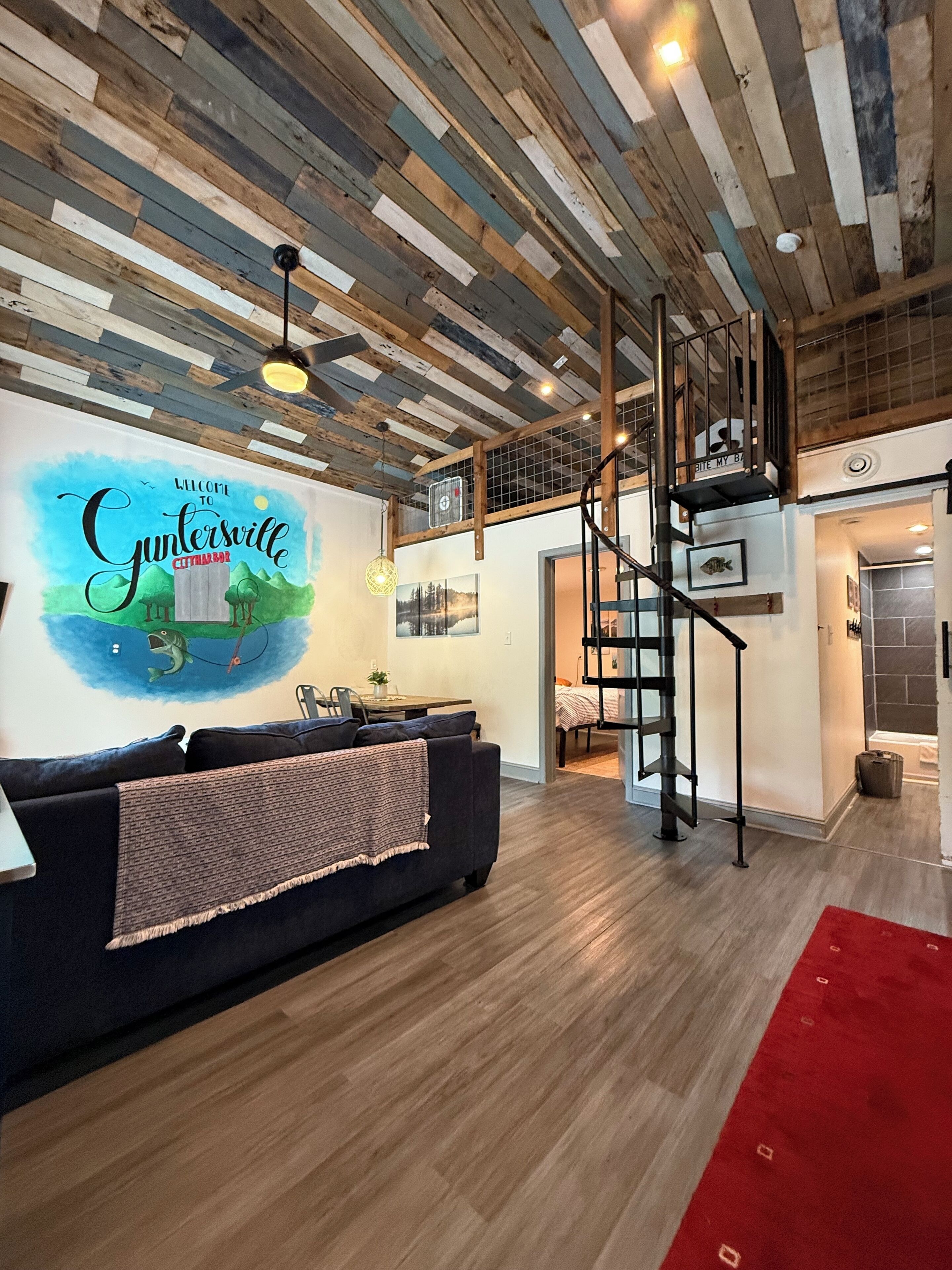 A modern living room with a dark sofa, wooden flooring, a red rug, and a staircase leading to an upper level.