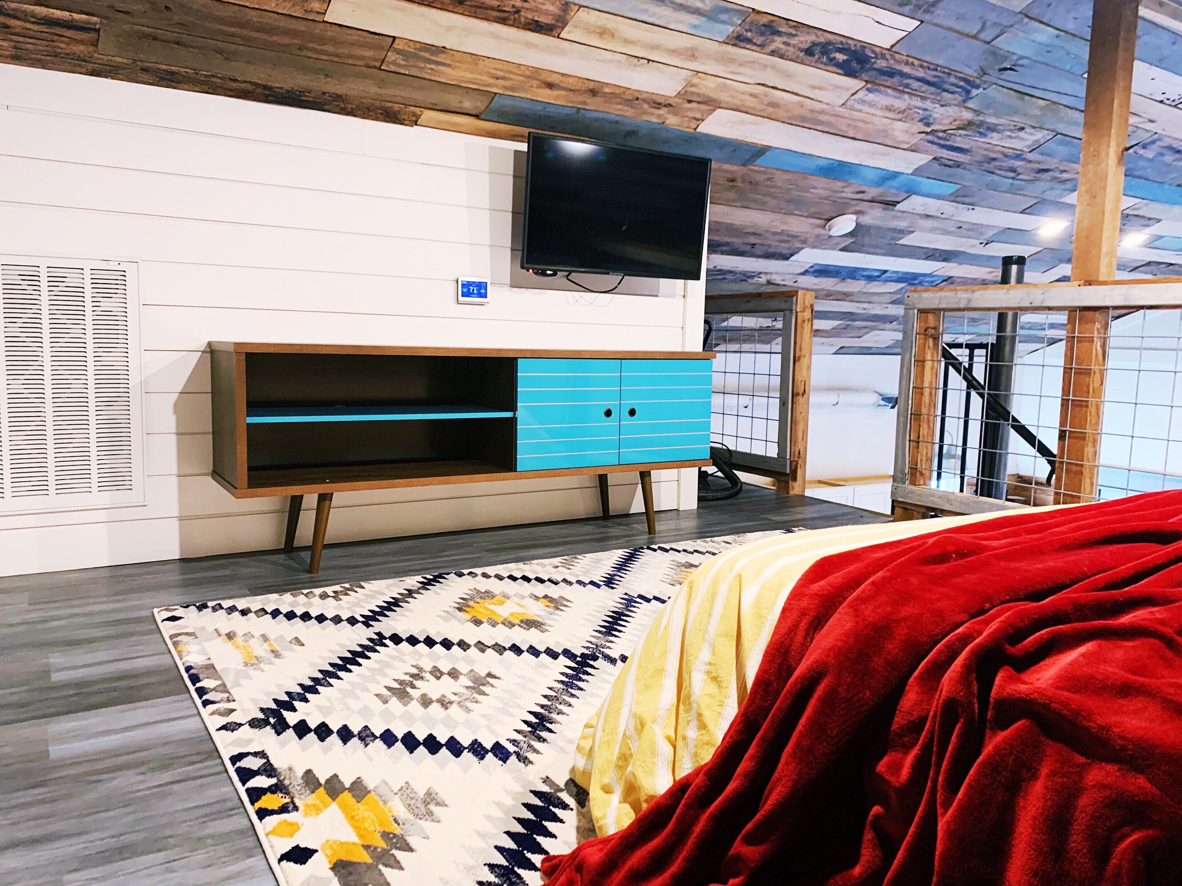 A bedroom with a TV mounted on the wall, a wooden cabinet with drawers, a patterned rug, and a red blanket on the bed.