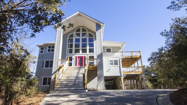 A two-story house with a prominent arched window, a front porch with a staircase, and a covered driveway. - Oceanside, Pool, Hot Tub, Close to Beach, OH1, Silver Sandcastle (Corolla)
