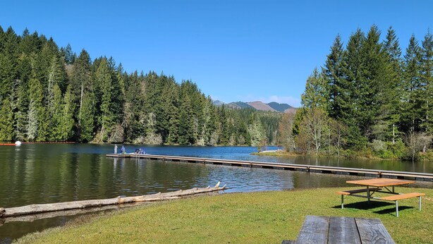 A lakeside view with a dock, picnic table, and forested hills in the background.