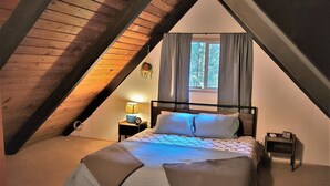A bedroom with a sloped ceiling, a bed with a patterned bedspread, a bedside lamp, and a window with curtains.