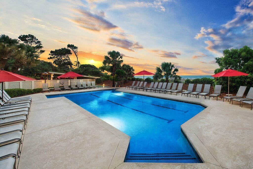 A swimming pool with lounge chairs and umbrellas, surrounded by trees and a sunset view.